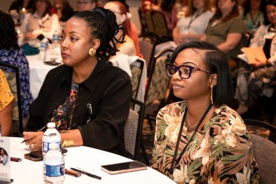 Two women sit at a table watching a speaker at a conference.