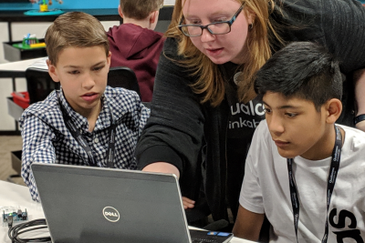 一位老师俯身在两个男孩的肩膀上，指着他们面前笔记本电脑屏幕上的某个东西。